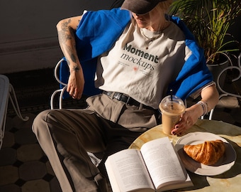 May include: A person wearing a brown baseball cap with the text "La Parisienne" and a white t-shirt with the text "Moment mockups" is sitting at a table with a book, a croissant, and a iced coffee.