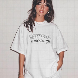 Puede incluir: Una camiseta blanca con el texto "Moment mockups" en negro, usada por una persona. La persona lleva pendientes de aro dorados y un collar dorado. El fondo es un patrón de cuadrícula blanca.