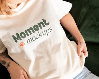 May include: A cream-coloured t-shirt with the text "Moment mockups" in dark green, and a small orange sunburst design. The shirt is being held up, showing the fabric's texture. The person is wearing a blue cap and light blue jeans.