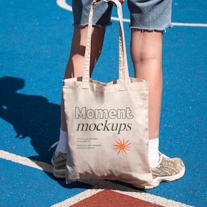 Op de afbeelding: Een witte canvas draagtas met de tekst "Moment mockups" en de slogan "This is your moment, come get it. Authentic moments for authentic brands." erop gedrukt. De tas wordt vastgehouden door een persoon op een blauw en rood oppervlak.