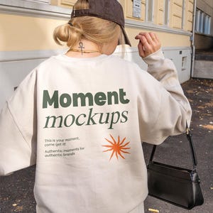 Może przedstawiać: Beżowa bluza z napisem "Moment mockups" w kolorze zielonym i pomarańczowym. Pod głównym tekstem znajduje się napis "This is your moment, come get it! Authentic moments for authentic brands". Po prawej stronie bluzy znajduje się stylizowany wzór słońca.