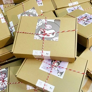 Puede incluir: Una pila de cajas de cart&oacute;n marr&oacute;n atadas con una cuerda roja y blanca. Cada caja tiene un dise&ntilde;o diferente con un personaje de dibujos animados con cabello negro y un lazo rosa. Las cajas est&aacute;n etiquetadas con el texto "Kuromi".