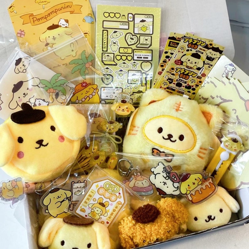 Puede incluir: Un peluche de Pompompurin amarillo y marr&oacute;n con un llavero de Pompompurin amarillo y marr&oacute;n. El peluche est&aacute; sentado en una caja con otros art&iacute;culos de Pompompurin, incluyendo pegatinas, llaveros y un peque&ntilde;o peluche. La caja est&aacute; llena de art&iacute;culos de Pompompurin amarillos y marrones.