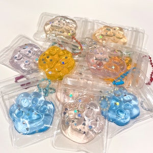 Könnte beinhalten: Eine Sammlung von bunten, glitzernden Pfotenabdruck-Schlüsselanhängern in durchsichtigen Plastiktüten. Die Pfotenabdrücke sind aus weichem, quietschweichem Material gefertigt und perfekt, um Ihren Schlüsseln oder Ihrer Tasche einen Hauch von Verspieltheit zu verleihen.