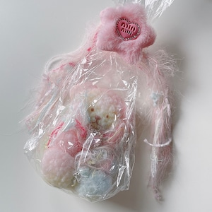 Puede incluir: Un oso de peluche rosa y blanco envuelto en plástico transparente con un amuleto en forma de corazón rosa unido al plástico.