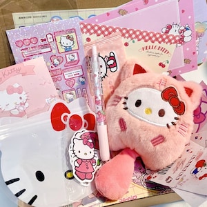 Puede incluir: Un peluche rosa de Hello Kitty con un lazo rojo y una cola a rayas rosa y blanca. El juguete est&aacute; rodeado de varios art&iacute;culos de papeler&iacute;a con tem&aacute;tica de Hello Kitty, incluidos pegatinas, blocs de notas y un bol&iacute;grafo.