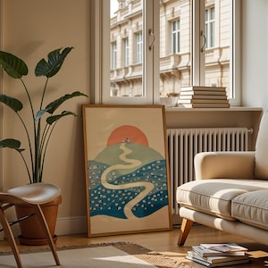 May include: A framed rectangular art print featuring a stylised landscape with a winding path leading towards a large orange sun over a green hill and a blue sea filled with small white shapes, resting on a wooden floor near a potted plant.