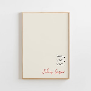 Puede incluir: Un póster enmarcado con fondo crema. El póster presenta la frase latina "Veni, vidi, vici" en texto negro, con "Julius Caesar" escrito en rojo debajo. El marco es de color madera clara.