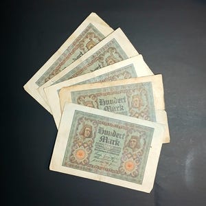 以下が含まれることがあります： 暗い背景に扇状に広げられた、いくつかのアンティークのドイツ100マルク紙幣。 各紙幣には肖像画、装飾的なボーダー、そして「Hundert Mark」の文字が描かれています。 紙幣は色あせ、古びた外観をしています。