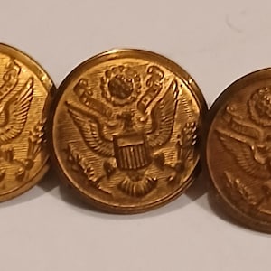 Może przedstawiać: Trzy złote metalowe guziki z szczegółowym wytłoczonym wzorem. Każdy guzik przedstawia orła z rozpostartymi skrzydłami, tarczą i innymi skomplikowanymi detalami. Guziki są okrągłe i wydają się być zabytkowe lub antyczne.