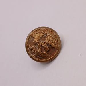 May include: A round, antique brass button with embossed details. The design includes a sunburst, a stylized building, a bird in flight, and the text "SNOWY FEET" on a banner. The button has a slightly worn appearance.