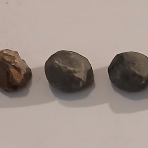 May include: Five small, irregularly shaped stones in varying shades of gray and brown are arranged in a row. The stones have a rough, textured surface, and are likely natural, possibly river rocks or similar. The stones are on a white surface.