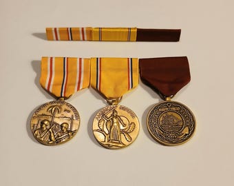 May include: Three vintage military medals with ribbons. The medals are gold-colored with detailed designs, including a ship, a woman with a sword, and soldiers on a beach. The ribbons are yellow, red, and brown.