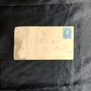 May include: A vintage rectangular paper envelope with a light tan, weathered appearance. A blue postage stamp featuring a portrait and the text Five Cents is affixed to the top right corner. Handwritten cursive text is visible on the envelope.