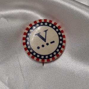 Puede incluir: Un botón redondo con un centro blanco que presenta una "V" azul y tres puntos, rodeado por un anillo de estrellas azules y rayas rojas y blancas. Se ve un alfiler en la parte posterior. Probablemente un artículo de campaña política.