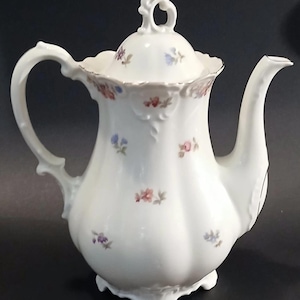 May include: White porcelain teapot with a curved handle and spout. The teapot features a lid with a decorative handle and is adorned with small floral patterns in shades of pink, blue, and purple. The rim is trimmed with gold.