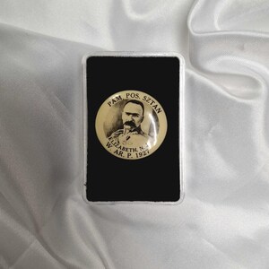 Pode incluir: Um alfinete comemorativo polonês vintage, apresentando um retrato em tons sépia de um homem com bigode. O alfinete está emoldurado em uma moldura retangular preta, com texto em polonês ao redor do retrato. O texto inclui as palavras "PAM. POS. SZTAN" e "ELIZABETH, N. W. AR. P. 1927".