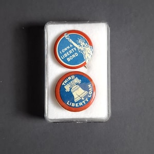 Pode incluir: Dois broches Liberty Bond vintage em uma caixa de plástico transparente. Um broche apresenta a Estátua da Liberdade e o texto "I Own a Liberty Bond". O outro broche tem um sino e diz "Third Liberty Loan".
