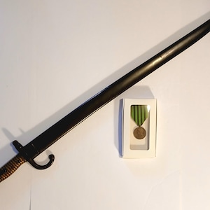 May include: A long, dark bayonet with a brass handle and a curved guard, alongside a medal in a white box. The bayonet is positioned diagonally, and the medal has a green and gold ribbon.