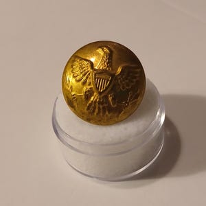 Może przedstawiać: Złoty guzik z wytłoczonym wzorem orła, umieszczony w przezroczystym plastikowym pojemniku. Guzik przedstawia szczegółowe przedstawienie orła z rozpostartymi skrzydłami, tarczą i gałązkami oliwnymi. Guzik jest okrągły i wydaje się być wykonany z metalu.