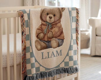 May include: A personalized baby blanket draped over a crib. The blanket features a brown teddy bear illustration wearing a brown jacket and blue pants. The name "LIAM" is printed in blue. The blanket has a blue and white checkered border and fringe.