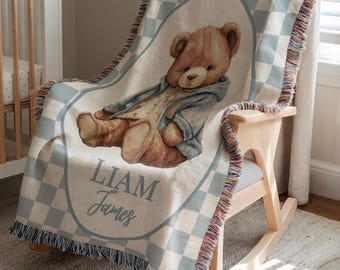 May include: A personalized baby blanket featuring a teddy bear illustration. The blanket has a light blue and white checkered pattern with a teddy bear in the center. The name "Liam James" is written in blue script. The blanket is draped over a wooden rocking chair.