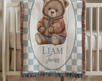 May include: A woven baby blanket featuring a watercolor illustration of a teddy bear wearing a hooded jacket and jeans. The blanket has a blue and white checkered border and the name "LIAM James" in script.