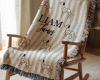May include: A gray and white striped throw blanket featuring dalmatian dog illustrations. The blanket has black fringe and the name "LIAM James" in black script. The blanket is draped over a wooden rocking chair.
