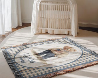May include: A woven baby blanket with a teddy bear design. The blanket features a blue and white gingham pattern border and a central oval with a teddy bear wearing a sweater with an American flag. The name "Liam James" is printed on the blanket.