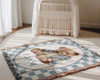 May include: A woven baby blanket featuring a watercolor illustration of a teddy bear wearing blue overalls. The blanket has a blue and white checkered border and the name "LIAM" is printed on the blanket.