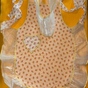 May include: A cream-coloured apron with a floral pattern and vertical stripes. It features a heart-shaped pocket and is trimmed with yellow and white ruffles. The straps and ties are patterned with small flowers, evoking a vintage aesthetic.
