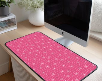 May include: A rectangular pink desk mat with a repeating pattern of white bows and dots on a wavy striped background. The mat has a black border and is placed on a wooden desk, with a computer monitor in the background.