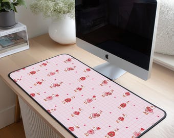 May include: A rectangular pink desk mat with a black border. The mat features a repeating pattern of cartoon geese holding hearts, set against a grid background. The desk mat is placed on a wooden desk.