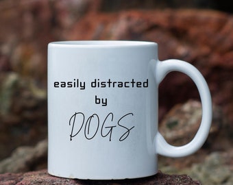 May include: White ceramic mug with black text that reads "easily distracted by DOGS". The mug is sitting on a pile of rocks.