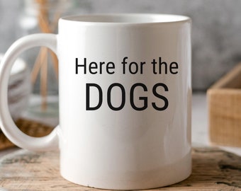 May include: White ceramic coffee mug with a black inscription that reads "Here for the DOGS". The mug is sitting on a wooden coaster.