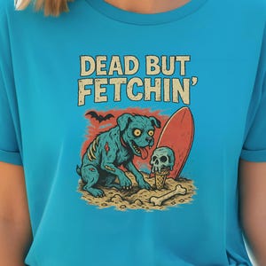 May include: Turquoise t-shirt with a graphic of a zombie dog, surfboard, skull, and bone on a beach. The text "DEAD BUT FETCHIN'" is above the graphic. The shirt has rolled-up sleeves.