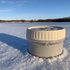 May include: A small, clear plastic container with a cream-colored lid sits on a snowy surface. The container holds a light-colored substance. The background features a frozen lake and a clear blue sky.
