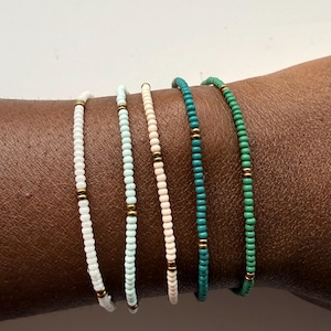 Puede incluir: Cinco pulseras de cuentas en blanco, azul claro, rosa, turquesa y verde. Cada pulsera presenta pequeñas cuentas de colores con detalles dorados. Las pulseras se muestran sobre un tono de piel oscuro, mostrando los colores y el diseño de la joyería.