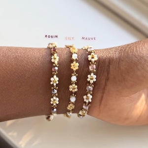 Op de afbeelding: Drie armbanden met bloemenontwerpen in wit, goud en paars worden getoond. De armbanden zijn gelabeld met "Robin", "Lily" en "Mauve". De armbanden worden om een pols gedragen.