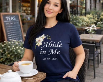 May include: A navy blue short-sleeve t-shirt featuring a graphic of white orchid flowers and the white text "Abide in Me" followed by "John 15:4" printed on the front. The shirt has a crew neckline and a standard fit.