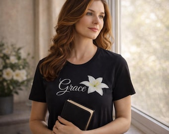 May include: Black t-shirt with the word "Grace" in cursive and a white lily graphic. The person in the image is holding a black book. The background includes a window and a floral arrangement.