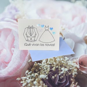 May include: A square wedding card with a tuxedo and wedding dress illustration, along with the text "¡Qué vivan los novios!" in black. The card has blue and silver heart accents and is set against a soft floral background.