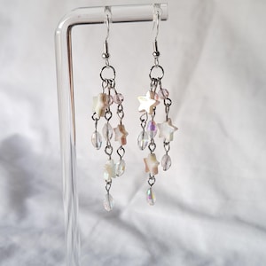 May include: Dangling earrings with a celestial theme. Each earring has a silver hook, a small chain, and multiple strands of iridescent beads and star-shaped charms. The earrings are displayed on a clear stand against a white backdrop.