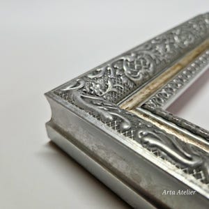 May include: Close-up of a silver picture frame with an ornate design. The frame has a raised, textured pattern and a slightly distressed finish. The frame is made of metal and has a white inner edge.