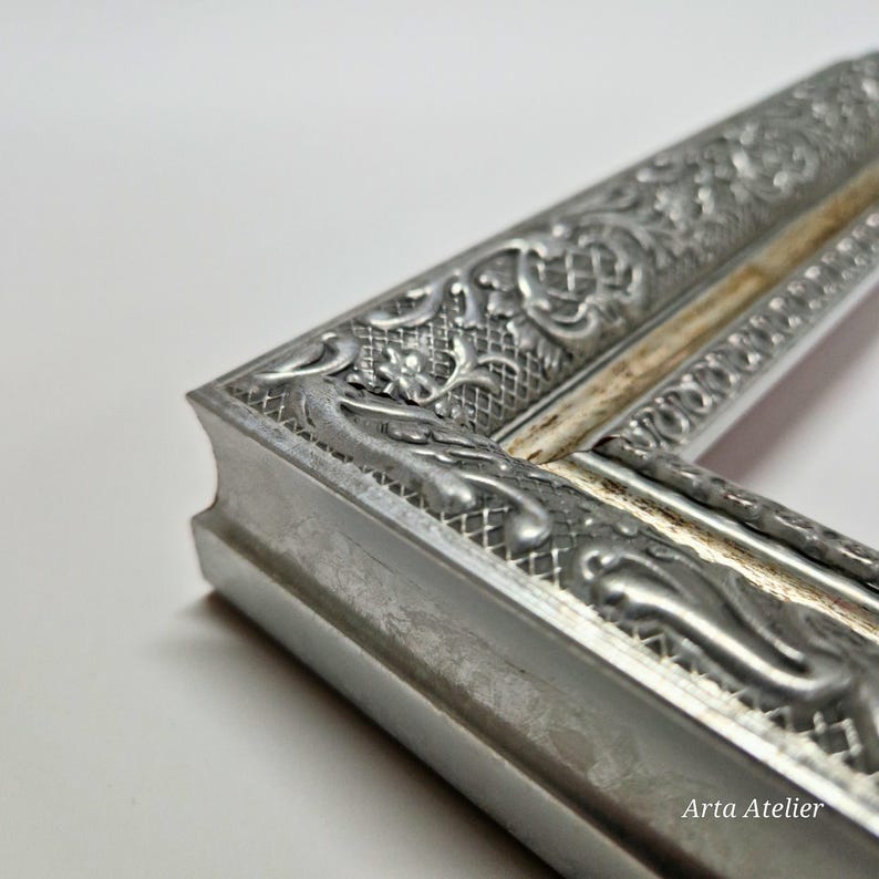May include: Close-up of a silver picture frame with an ornate design. The frame has a raised, textured pattern and a slightly distressed finish. The frame is made of metal and has a white inner edge.