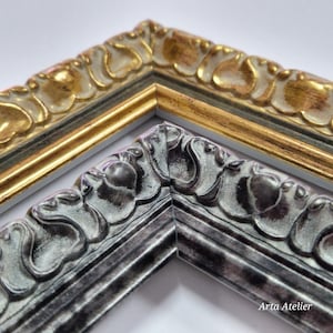 May include: Close-up of two ornate picture frame corners. One is gold with a cream-colored floral design, and the other is silver with a similar pattern. The frames are angled, showcasing their detailed craftsmanship and decorative style.