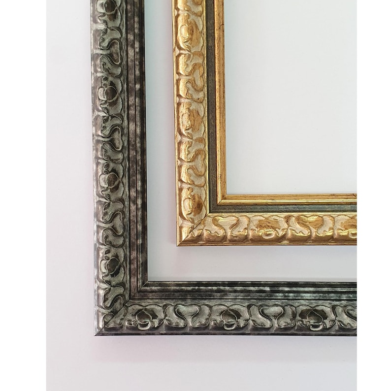 May include: Two ornate picture frames. One frame is silver with a dark gray wash, and the other is gold with a dark green wash. Both frames have a detailed, repeating floral pattern. The frames are empty, with a white background.