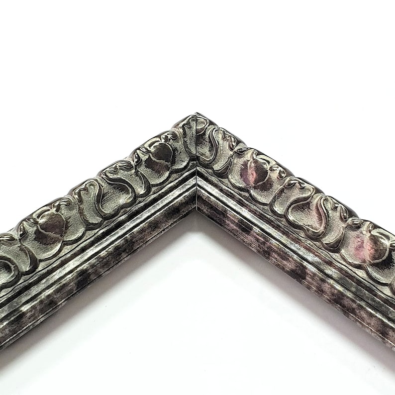 May include: Close-up of a corner of a silver picture frame with an ornate, detailed design. The frame has a distressed, aged look.