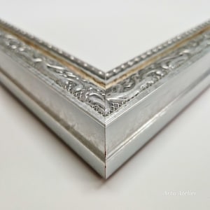 May include: Close-up of the corner of a silver picture frame with an ornate design. The frame has a raised edge and a smooth, polished finish.