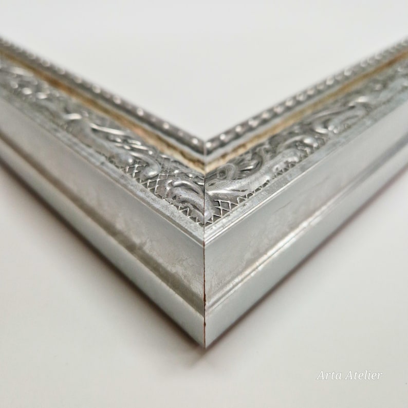 May include: Close-up of the corner of a silver picture frame with an ornate design. The frame has a raised edge and a smooth, polished finish.